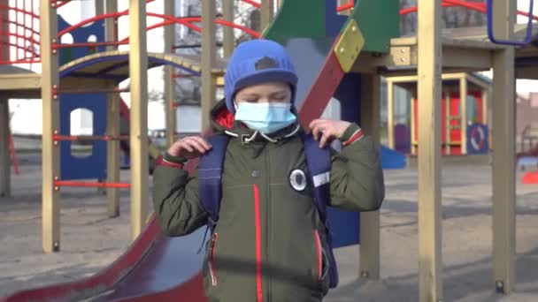 Yüzü maskeli beyaz okul çocuğu çocuk parkında tek başına duruyor. Sırt çantalı küçük şirin çocuğun portresi ilkbahar gününü dışarıda Covid-19 'da kilitli geçiriyor. Coronavirus karantinası, virüs. — Stok video