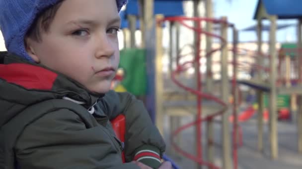 Vista lateral close-up do triste menino caucasiano sozinho sentado no parque infantil no dia de primavera durante o bloqueio do coronavírus. O estudante passa o tempo livre ao ar livre. Covid-19, quarentena, lazer, estilo de vida . — Vídeo de Stock