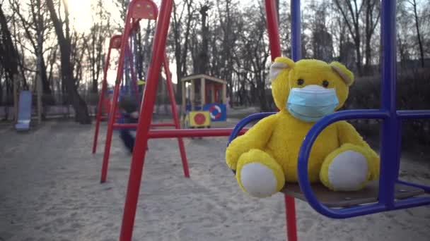 Close-up van gele teddybeer in gezichtsmasker op kinderspeelplaats met jongen op schommel. Vrijetijdsbesteding buiten op coronavirus quarantaine. Gezondheidszorg, Covid-19 afsluiting, wereldwijde pandemie. — Stockvideo