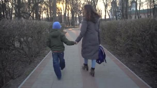 Achteraanzicht van de jonge brunette Kaukasische vrouw en kleine schooljongen wandelen 's avonds door het steegje in het stadspark. Moeder en zoon keren terug naar huis na het werk en de lessen. Levensstijl. — Stockvideo