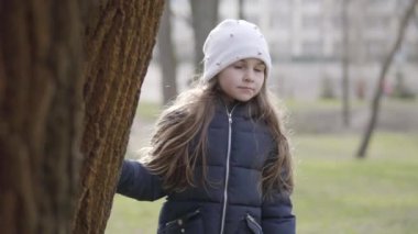 Güzel küçük kız ağacın yanında park yerinde duruyor ve başka tarafa bakıyor. Güneşli baharda ya da sonbahar günü açık havada dinlenen beyaz bir çocuğun portresi. Boş zaman, çocukluk, yaşam tarzı.