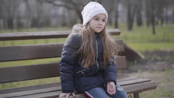 Brunette schoolmeisje zit op een bankje in het park en heft haar been op. Portret van een eenzaam sportief kind dat buiten rust op zonnige lente- of herfstdag. Vrije tijd, levensstijl, eenzaamheid. — Stockvideo