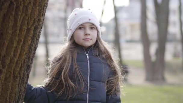 Porträt eines überraschten kaukasischen Mädchens, das in die Kamera blickt und sich hinter dem Baumstamm versteckt. Fröhliches Kind, das sich an einem sonnigen Frühlingstag im Freien vergnügt. Natur, Lebensstil, Entspannung. — Stockvideo