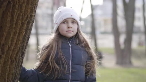 Retrato de niña caucásica sorprendida mirando a la cámara y escondida detrás del tronco del árbol. Chico alegre divirtiéndose al aire libre en el soleado día de primavera. Naturaleza, estilo de vida, relajación . — Vídeos de Stock