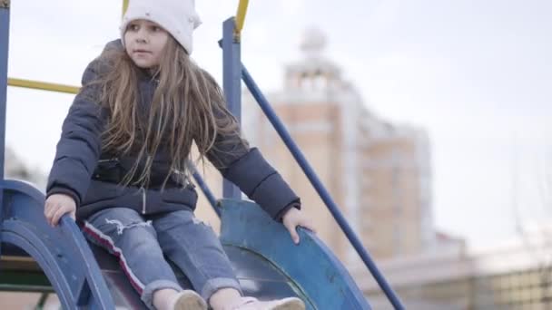 En glad liten flicka glider ner för rutschkanan på lekplatsen. Kamera efter kaukasiska barn har roligt utomhus på solig dag. Fritid, livsstil, glädje, nöje, barndom. — Stockvideo