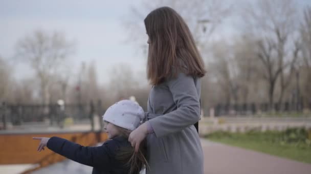 Vista lateral de la madre y la hija caucásicas felices de pie en el parque y hablando. Amistosa familia pasar tiempo libre en el parque en el día soleado. Estilo de vida, unidad, ocio, felicidad . — Vídeos de Stock