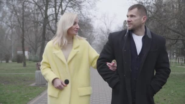 Porträtt av unga kaukasiska man och kvinna chattar och ler som promenader längs gränden i parken. Positivt par njuter av molnig dag utomhus. Fritid, livsstil, lycka, dejting. — Stockvideo