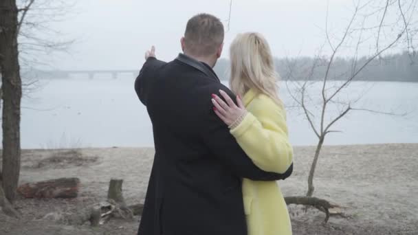 Back view of confident brunette man hugging blond woman, pointing away and talking. Elegant husband dating with loving wife on riverbank. Joy, leisure, romance, love, lifestyle. — Stock Video