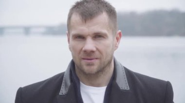 Gri gözlü, açık havada poz veren esmer, esmer, esmer bir adamın yakın çekimi. Bulutlu bir günde nehir kıyısında duran zarif ceketli genç adamın portresi. Yaşam tarzı, güven, rahatlık..