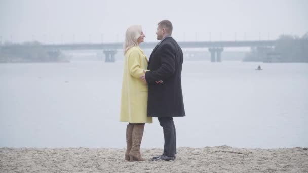 Mujer rubia caucásica abrazando a un hombre cariñoso en la orilla del río. Vista lateral amplia toma de pareja casada citas al aire libre. Amor, matrimonio, relación, estilo de vida . — Vídeo de stock