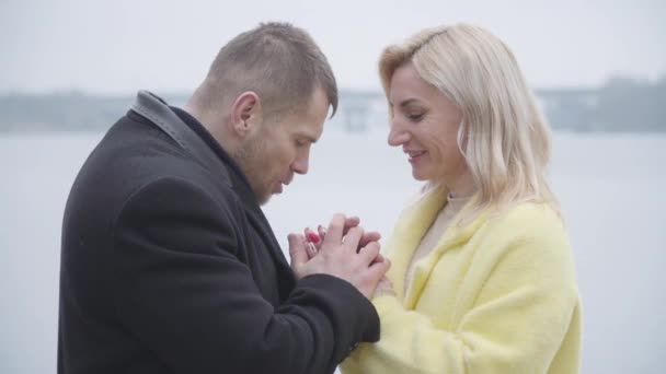 Side view of Caucasian man in love warming hands of blond woman with breathe. Loving guy taking care of sweetheart as standing on riverbank. Joy, leisure, lifestyle, romance, bonding. — Stock Video