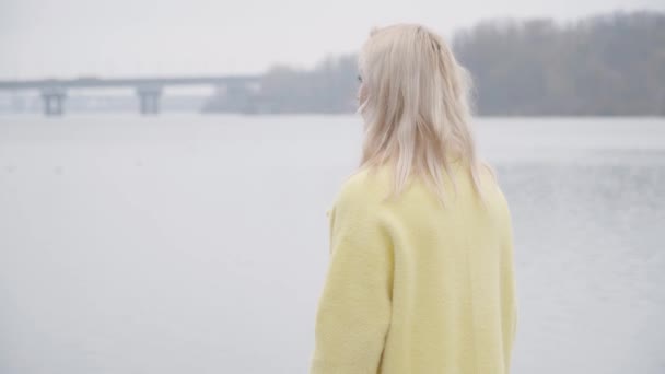 Femme blanche blonde solitaire en manteau jaune debout sur la rive et regardant loin. Vue de face arrière de belle dame profitant d'une journée calme et nuageuse en plein air. Loisirs, mode de vie, tranquillité . — Video