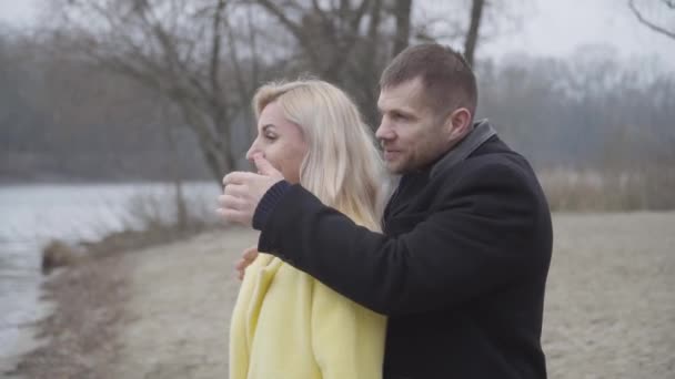 Positivo hombre caucásico abriendo los ojos de la mujer rubia sonriente en la orilla del río y besándose cariño. Pareja feliz disfrutando de citas al aire libre. Amor, romance, estilo de vida . — Vídeo de stock