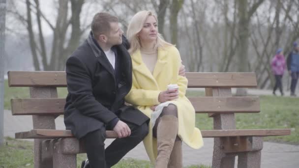 Romantic Caucasian couple sitting on bench in park, talking and rubbing noses. Portrait of loving adult man and woman dating outdoors on spring or autumn day. Happiness, romance, leisure. — Stock Video