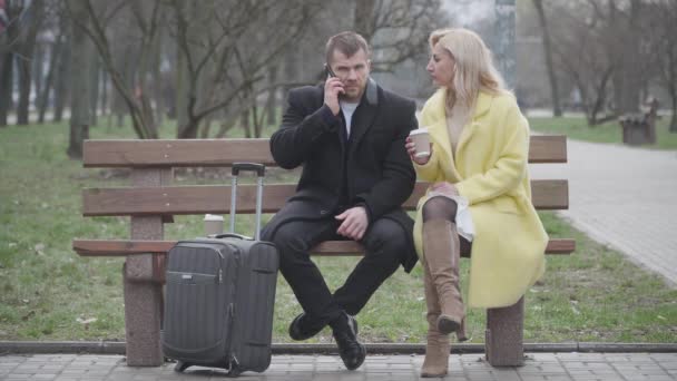 Porträt eines ernsthaften erwachsenen kaukasischen Paares mit Reisetasche, das im Park auf ein Taxi wartet. Mann telefoniert mit Frau auf Bank mit Kaffeetasse. Tourismus, Lebensstil, Reisen, Reisen. — Stockvideo