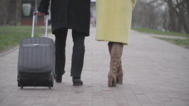 Dişi ve erkek bacaklarının arka görüntüsü, seyahat çantasıyla sokakta yürüyorlar. Tanımlanamayan rahat kadın ve erkek yabancı bir şehirde parkta geziniyorlar. Turizm, seyahat, eğlence, yaşam tarzı.