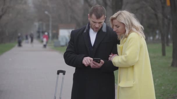 Pareja de adultos caucásicos de pie en el parque con la maleta y el uso de teléfono inteligente. Turistas masculinos y femeninos deciden sobre el destino turístico en la ciudad extranjera. Ocio, turismo, viajes, estilo de vida . — Vídeo de stock