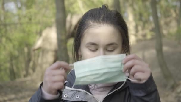 Серйозна молода жінка одягає маску обличчя і дивиться на камеру. Знятий портрет сіроокої кавказької дівчини, яка позувала в сонячному парку на карантині Ковід-19. Локдаун, спосіб життя, пандемія. — стокове відео