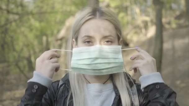 Mujer rubia caucásica poniéndose mascarilla y mirando a la cámara. Cara de cerca de la joven seria y confiada posando en el parque en el soleado día de primavera. Covid-19, coronavirus, cuarentena, bloqueo . — Vídeo de stock