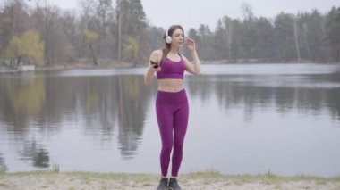 Parktaki göl kıyısında müzik dinleyen ve dans eden mor spor giysili esmer kadın. Neşeli Kafkasyalı sporcu kadın sabah sporunun tadını çıkarıyor..