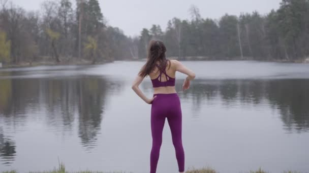Widok z tyłu szczupłej białej brunetki rozciągającej się na brzegu jeziora. Ćwiczenie pewnej siebie szczupłej kobiety na świeżym powietrzu. Sport, zdrowy styl życia, piękno, natura. — Wideo stockowe