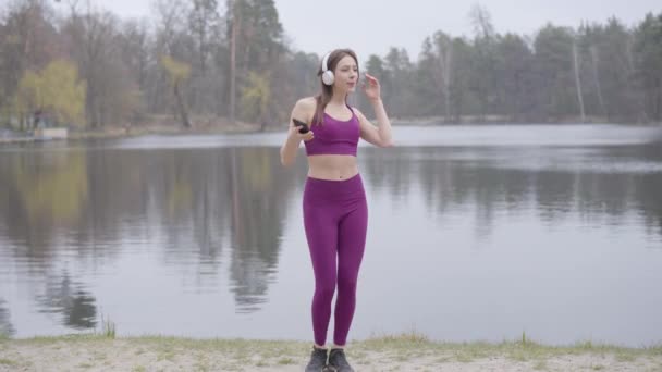 Parktaki göl kıyısında müzik dinleyen ve dans eden mor spor giysili esmer kadın. Neşeli Kafkasyalı sporcu kadın sabah sporunun tadını çıkarıyor.. — Stok video