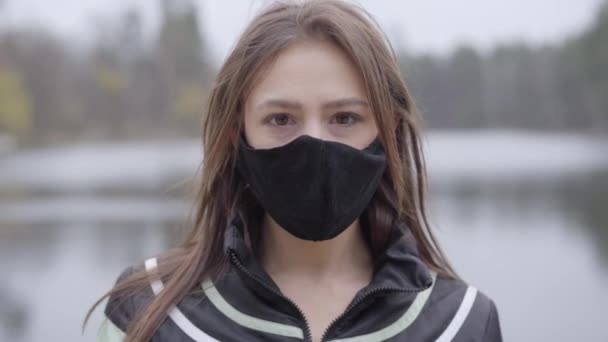 Primer plano retrato de morena de ojos marrones mujer caucásica en máscara protectora mirando a la cámara. Hermosa chica de pie en la orilla del lago en el día nublado. Salud, Covid-19 cuarentena, estilo de vida . — Vídeo de stock