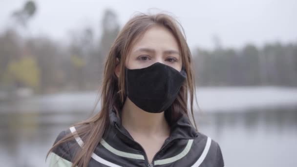 Charmante brune jeune femme qui tourne le visage vers la caméra et enlève le masque protecteur. Portrait d'heureuse fille aux yeux bruns profitant de la fin de la quarantaine de coronavirus à l'extérieur. Concept de pandémie Covid-19 . — Video