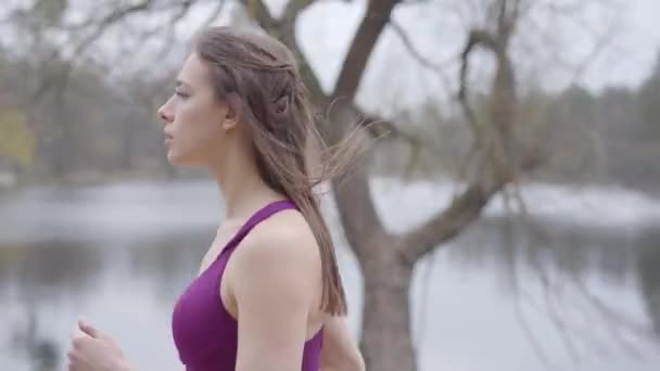 Boční pohled na sebevědomou bělošku běžící podél břehu jezera v parku. Štíhlá brunetka ráno běhá venku. Fitness, zdravý životní styl, sport, cvičení. — Stock video