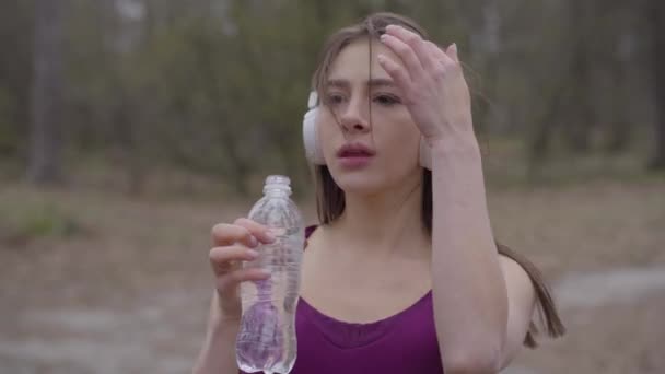 Portret van de vrolijke jonge Kaukasische sportvrouw in koptelefoon die water drinkt tijdens het sporten buiten. Lachende mooie meid training in het park in de ochtend. Vreugde, gezondheid, fitness, lifetsyle. — Stockvideo