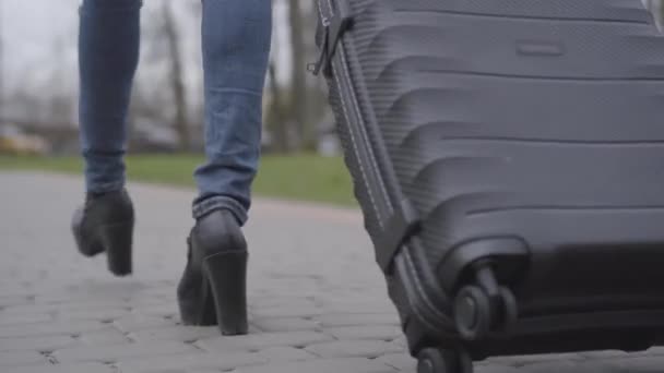 Fotocamera che segue le gambe femminili in tacchi alti e jeans passeggiando lungo il vicolo del parco con borsa da viaggio. Donna magra irriconoscibile che viaggia con valigia all'aperto. Tempo libero, stile di vita, turismo . — Video Stock