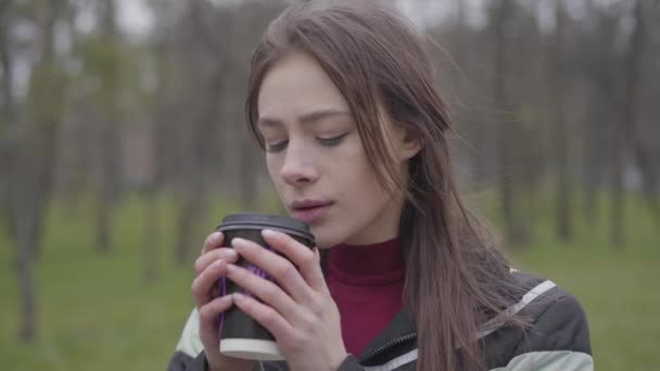 Zbliżenie troskliwej, czarującej dziewczyny pijącej kawę na świeżym powietrzu. Portret pięknej brunetki z brązowymi oczami cieszącej się pochmurnym dniem w parku. Styl życia, wypoczynek, odpoczynek, natura. — Wideo stockowe