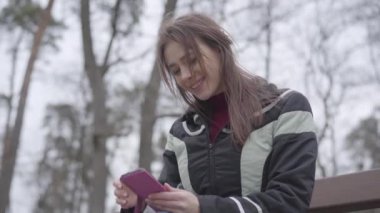 Akıllı telefon kullanan mutlu güzel bir kızın portresi ve gülüşü parkta bankta oturuyormuş gibi. Genç esmer kadın açık havada bulutlu bir sabah geçiriyor. Eğlence, yaşam tarzı, eğlence, mutluluk..