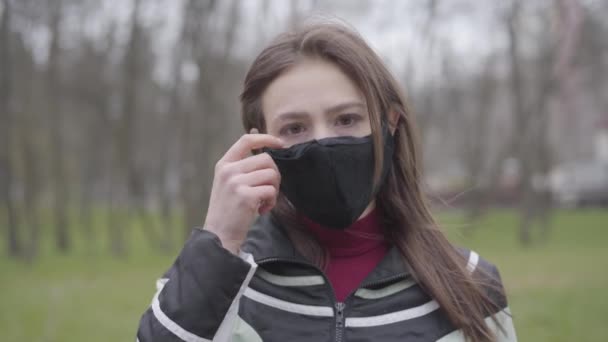 Primer plano de la alegre joven mujer caucásica quitándose la mascarilla y sonriendo a la cámara. Chica feliz disfrutando de la terminación de la cuarentena del coronavirus al aire libre. Covid-19 pandemia final . — Vídeos de Stock