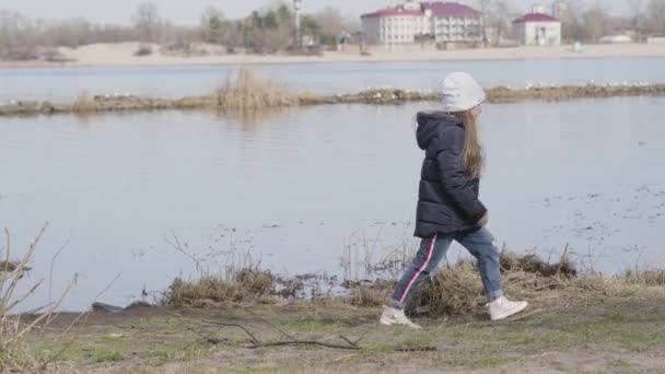 Široký záběr malé bělošky procházející se podél břehu řeky nebo jezera s roztomilým malým pinčourem a odcházející. Šťastné dítě a mazlíček odpočívá venku za slunečného jarního dne. Volný čas, jednota, životní styl. — Stock video