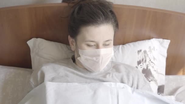 Draufsicht einer jungen kranken Frau, die niest und hustet. Porträt eines kaukasischen brünetten Mädchens mit grauen Augen und Gesichtsmaske, das auf der Krankenstation im Bett der Covid-19-Pandemie liegt. S-log-2 ProRes 422 HQ. — Stockvideo