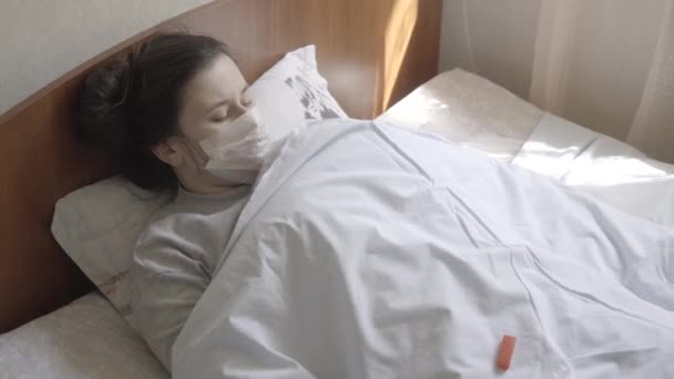Üzgün Kafkas esmer kadın vücut ısısını termometreyle ölçüyor. Covid-19 hastalığından muzdarip, yüzü maskeli mutsuz bir beyaz kız yatakta yatıyor. S-log-2 ProRes 422 Merkez. — Stok video