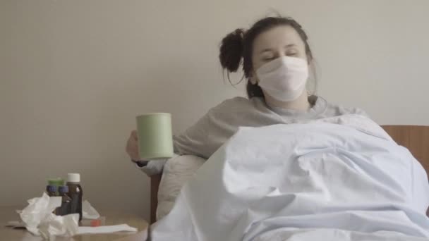 一个疲惫的白种人女人戴着面具躺在床上喝茶的画像。年轻疲惫的黑发女孩患有Covid-19流行病。救护车治疗，疾病。S-log-2 ProRes 422总部. — 图库视频影像