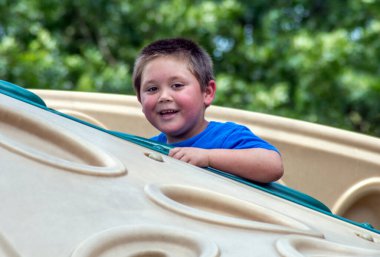 Gülümseyen küçük çocuk, açık hava parkında ve oyun alanında yüksek etkiye sahip bir oyun yapısına tırmanıyor.