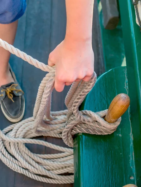 A sailor secures ropes from a sail of a tall ship, around a wooden post on the side of this vessel