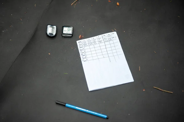 Two bike speedometers of cyclists lying on the needles grey sleeping mat pad in coniferous forest with pen and notebook. Close up. Wandering on bikes. Bikepacking, Sumava, Bohemian Forest, Bohmerwald, Czech Republic.