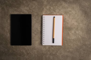 Kahverengi arka planda siyah tablet, not defteri ve kalem.