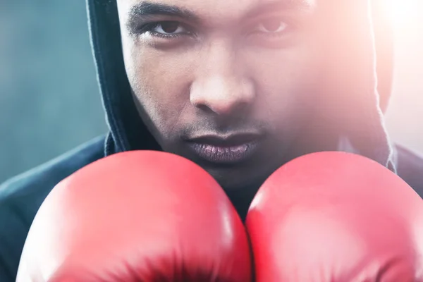 Gros plan du boxeur afro-américain portant des gants rouges et un sweat à capuche noir et debout sur fond gris. Concept de carrière sportive — Photo