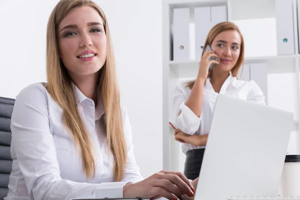 Två affärskvinnor i ämbetet. Man skriver på sitt bärbara tangentbord. Den andra pratar på telefon och tittar på sin kollega. Konceptet att arbeta tillsammans. — Stockfoto