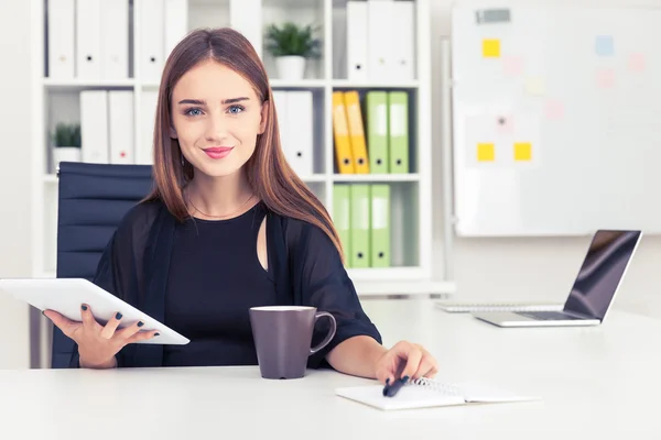 Smilinig affärskvinna håller sin penna och Tablet PC och sitter i vitt kontor med färgade mappar i bokhyllan — Stockfoto