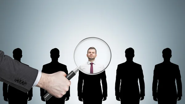 Large human hand with magnifying glass examining man silhouettes against light gray wall. Concept of recruitment. Mockup — Stock Photo, Image