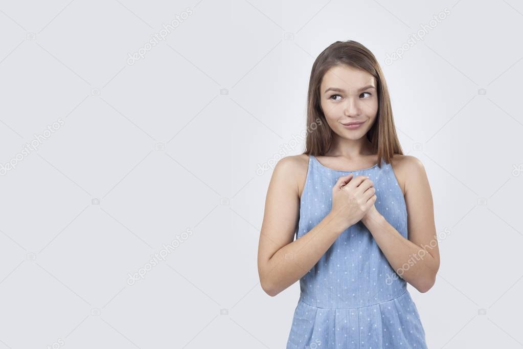 Shy girl in blue dress is standing against gray background