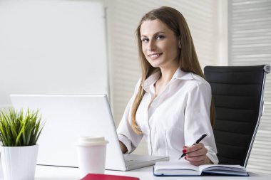 Beyaz bir bluz giyen ve ile bir dizüstü bilgisayar masasında oturan gülümseyen ve güzel bir işkadını, yakın çekim.