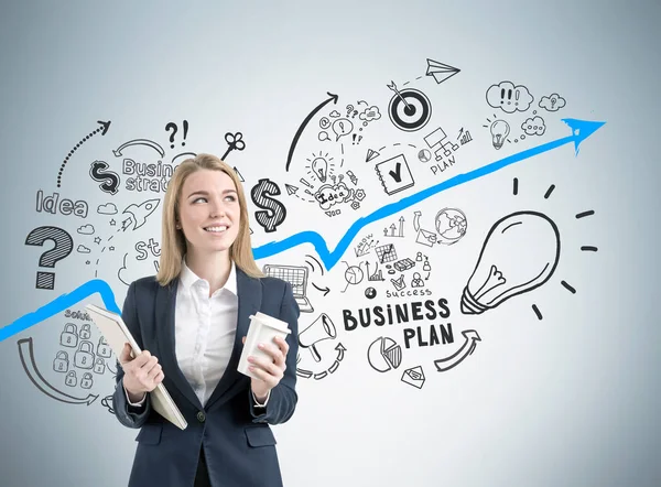 Smiling blond woman holding a notebook and a paper cup of coffee is standing near a gray wall with a blue graph and business plan icons. — Stock Photo, Image