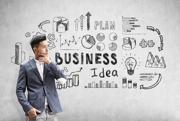 Pensativo empresario asiático está de pie cerca de una pared de hormigón con negro boceto idea de negocio representado en él . —  Fotos de Stock